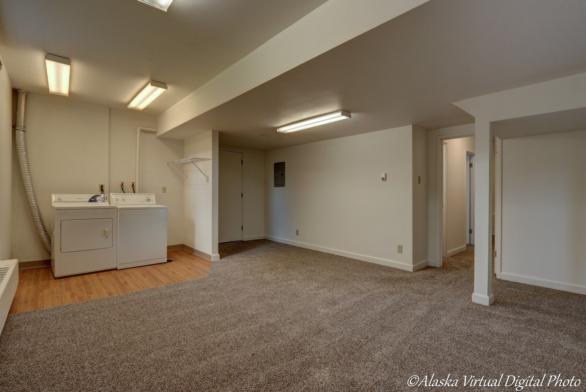 Image of large basement area with fluorescent lighting and carpeted floor. Small nook with wood floor houses washer and dryer